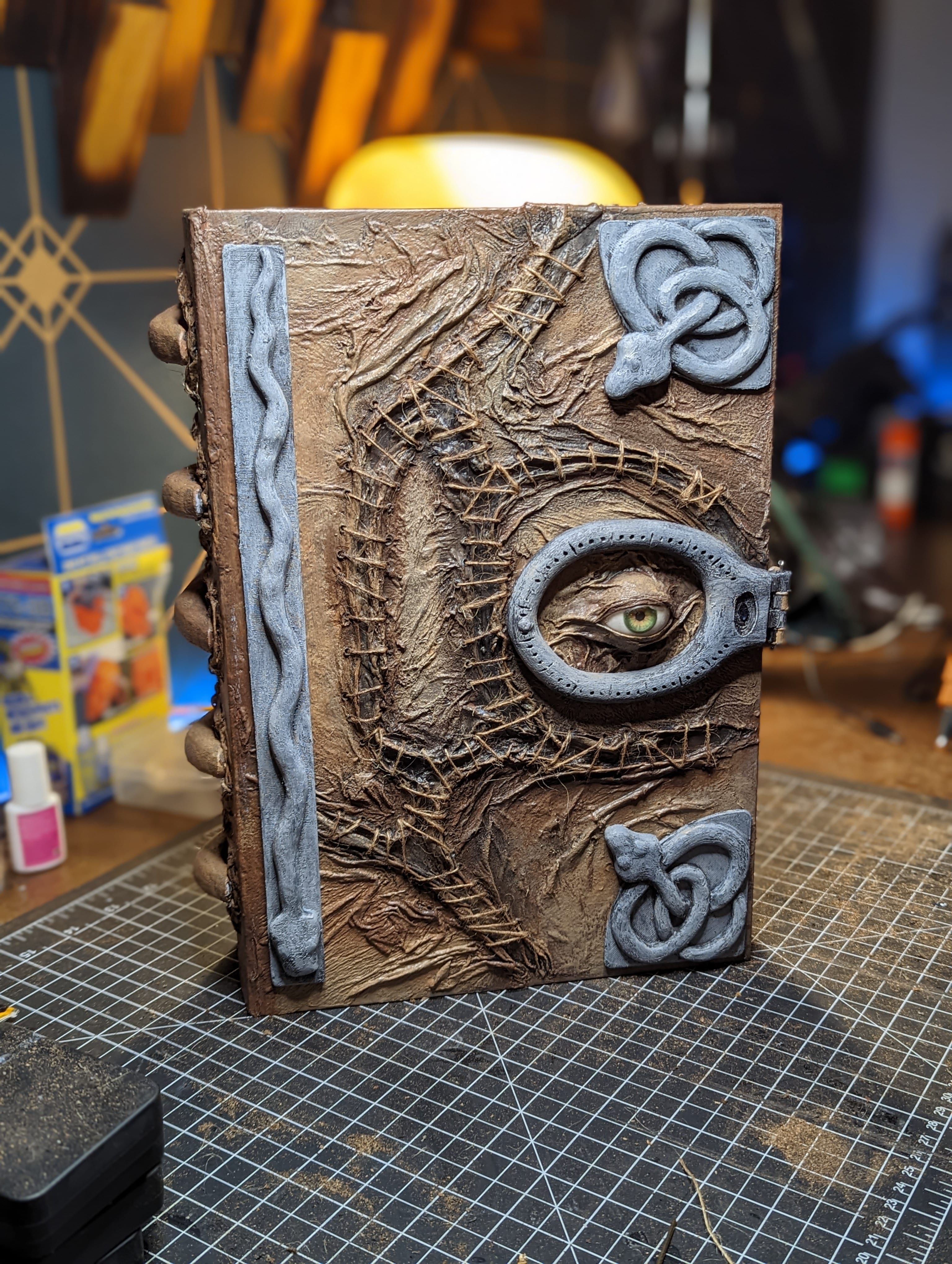 The finished Book standing upright on the workbench, with a green eye, twine-stitched skin, and painted gray hardware across its front cover.