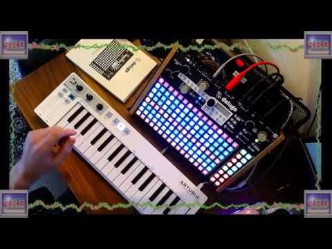 An overhead view of a hand playing a small white keyboard beside a lit Deluge and another synthesizer.