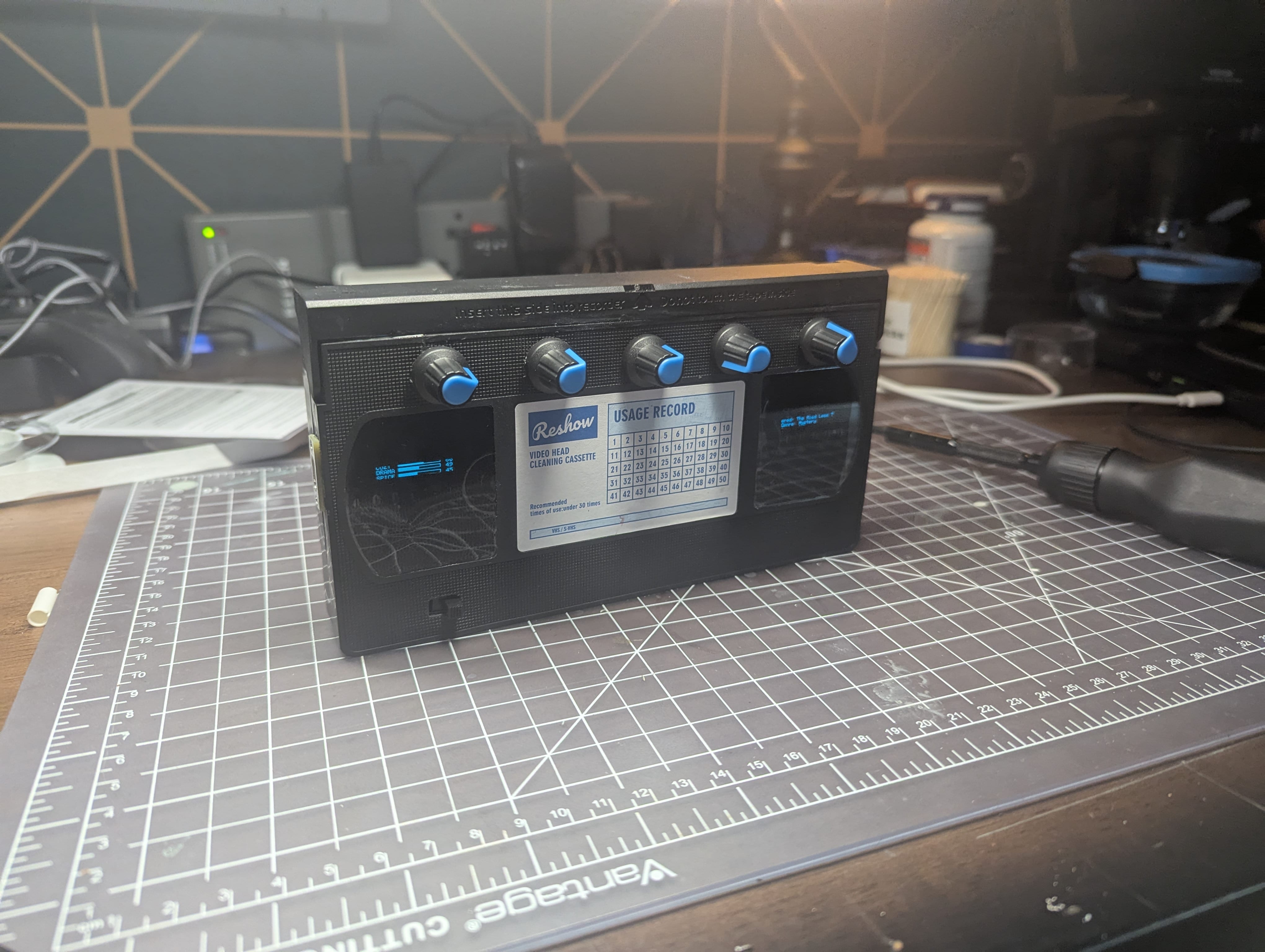 The powered Halmarker standing on a workbench, with five blue-tipped dials above two OLED screens set into a VHS head-cleaning cassette.