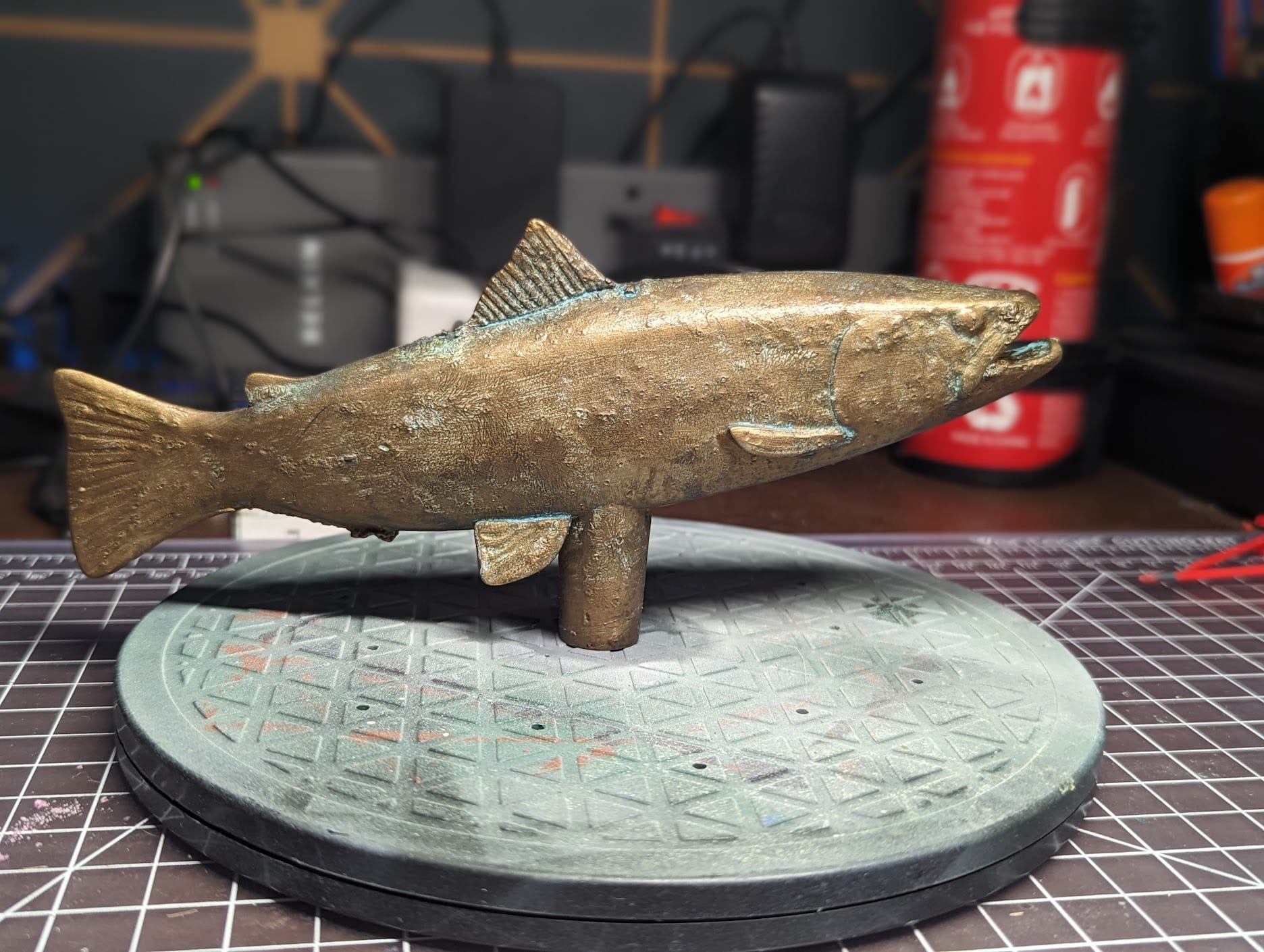 The finished brown trout finial in profile on a workbench, its bronze-and-brass surface darkened with patina.