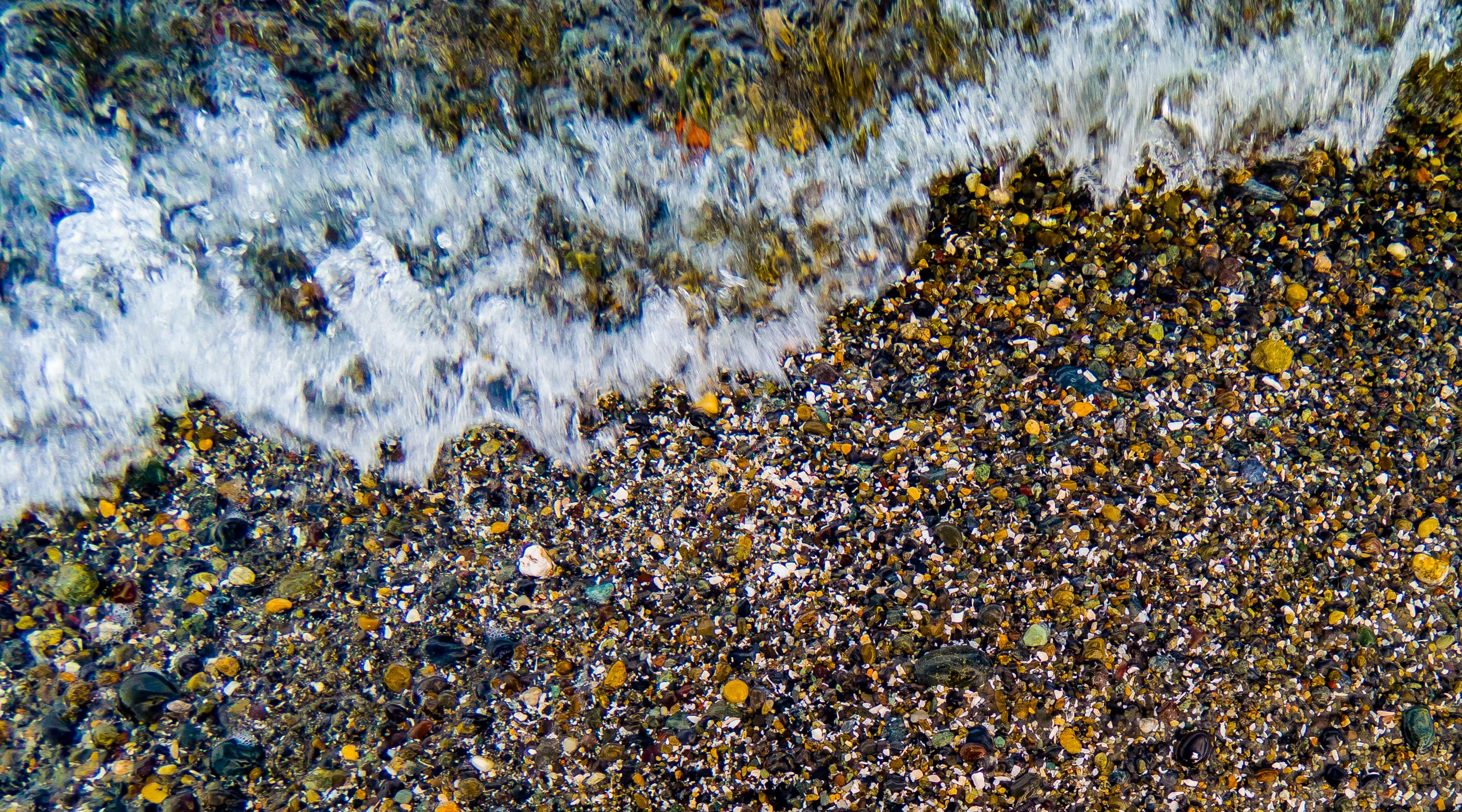 A foamy wave washes over multicolored pebbles on the shoreline.