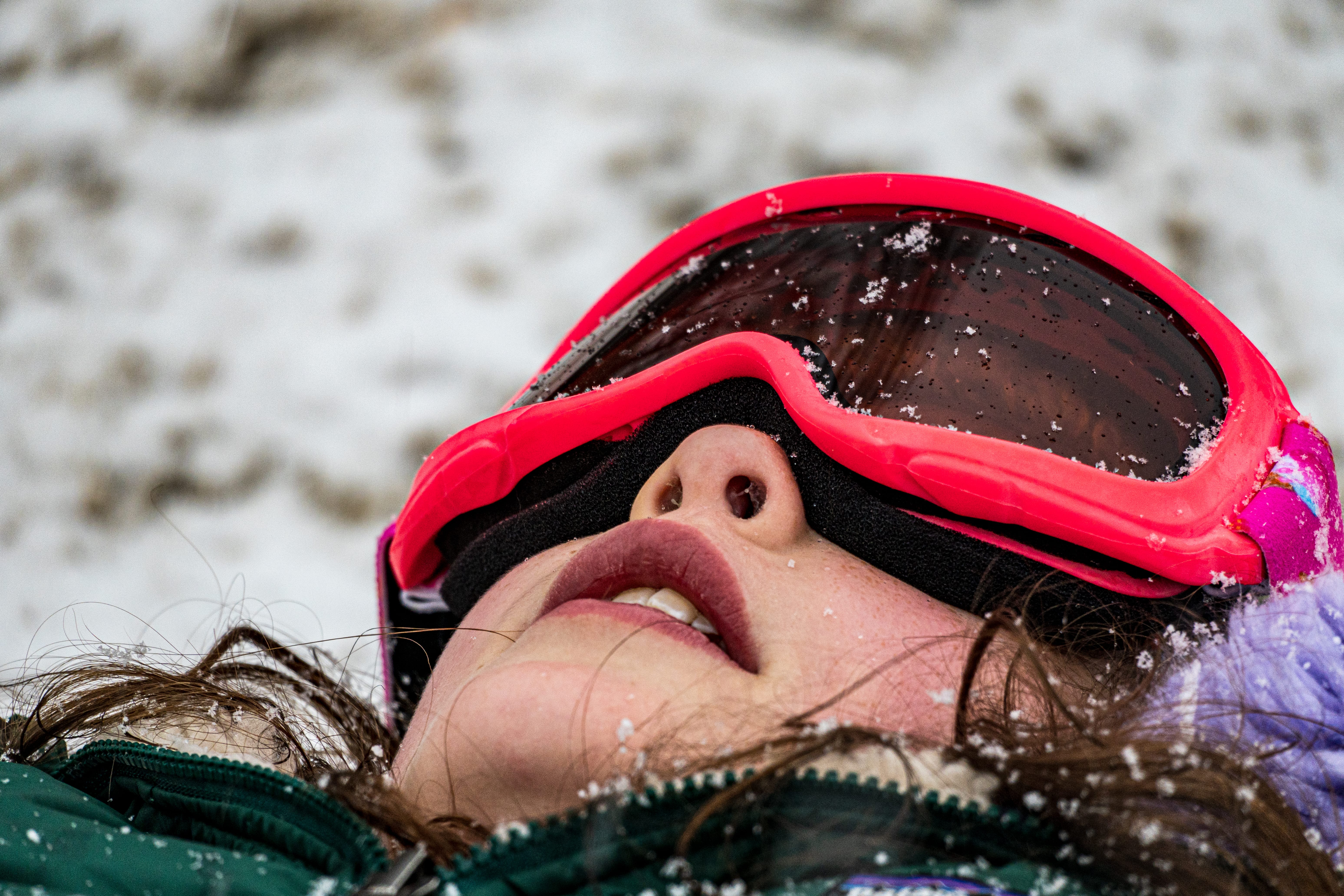 A child in red snow goggles lies on their back in fresh snow.