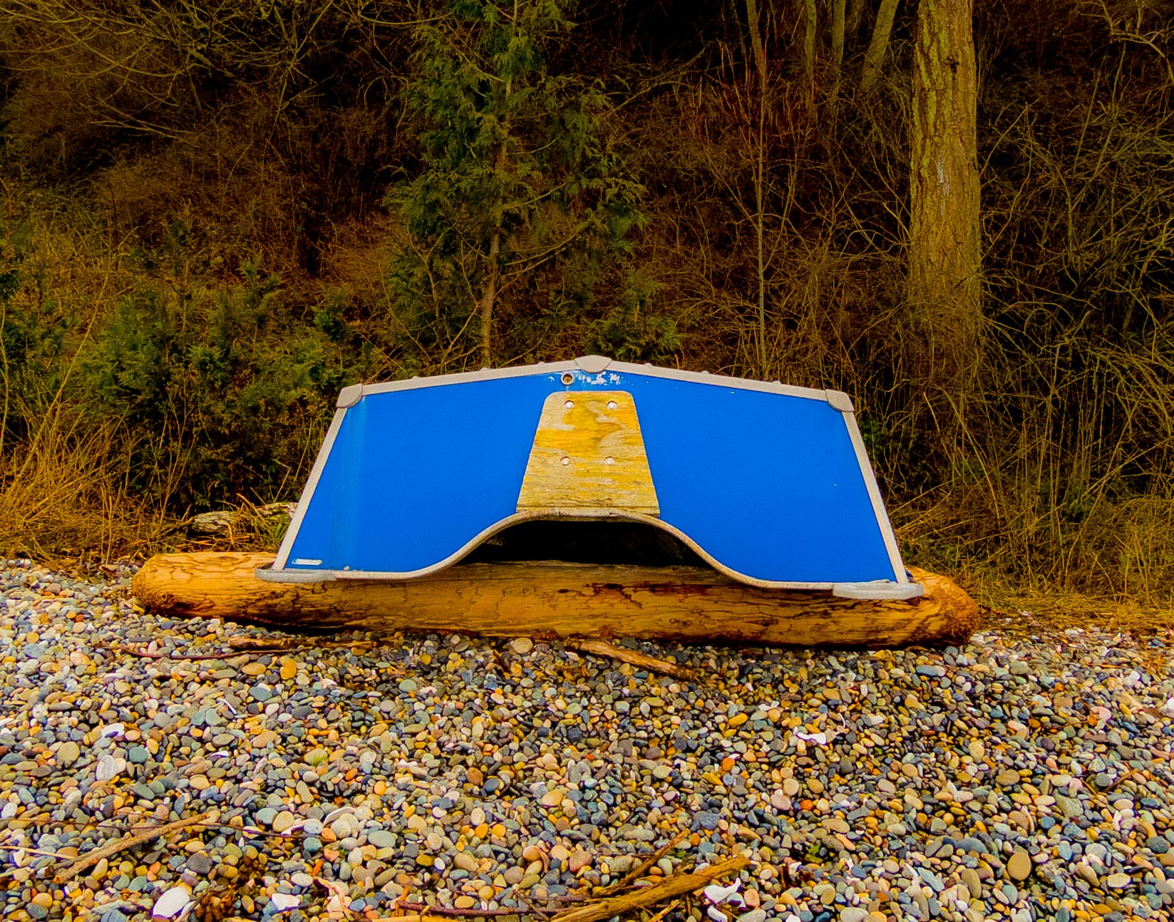 An overturned blue-and-white rowboat rests on a pebble beach at the edge of the woods.