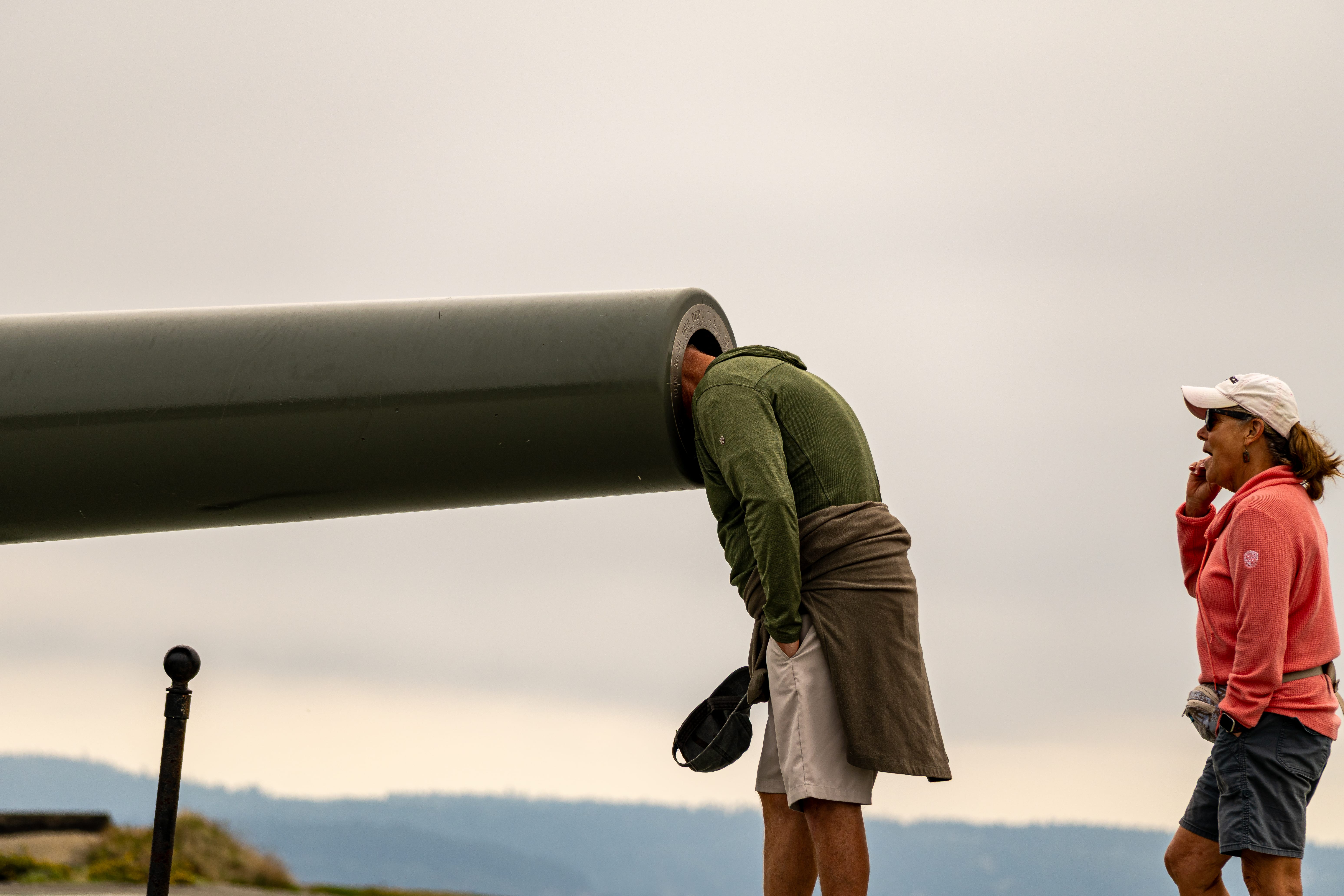 Two visitors examine a large green viewing tube beneath an overcast sky.