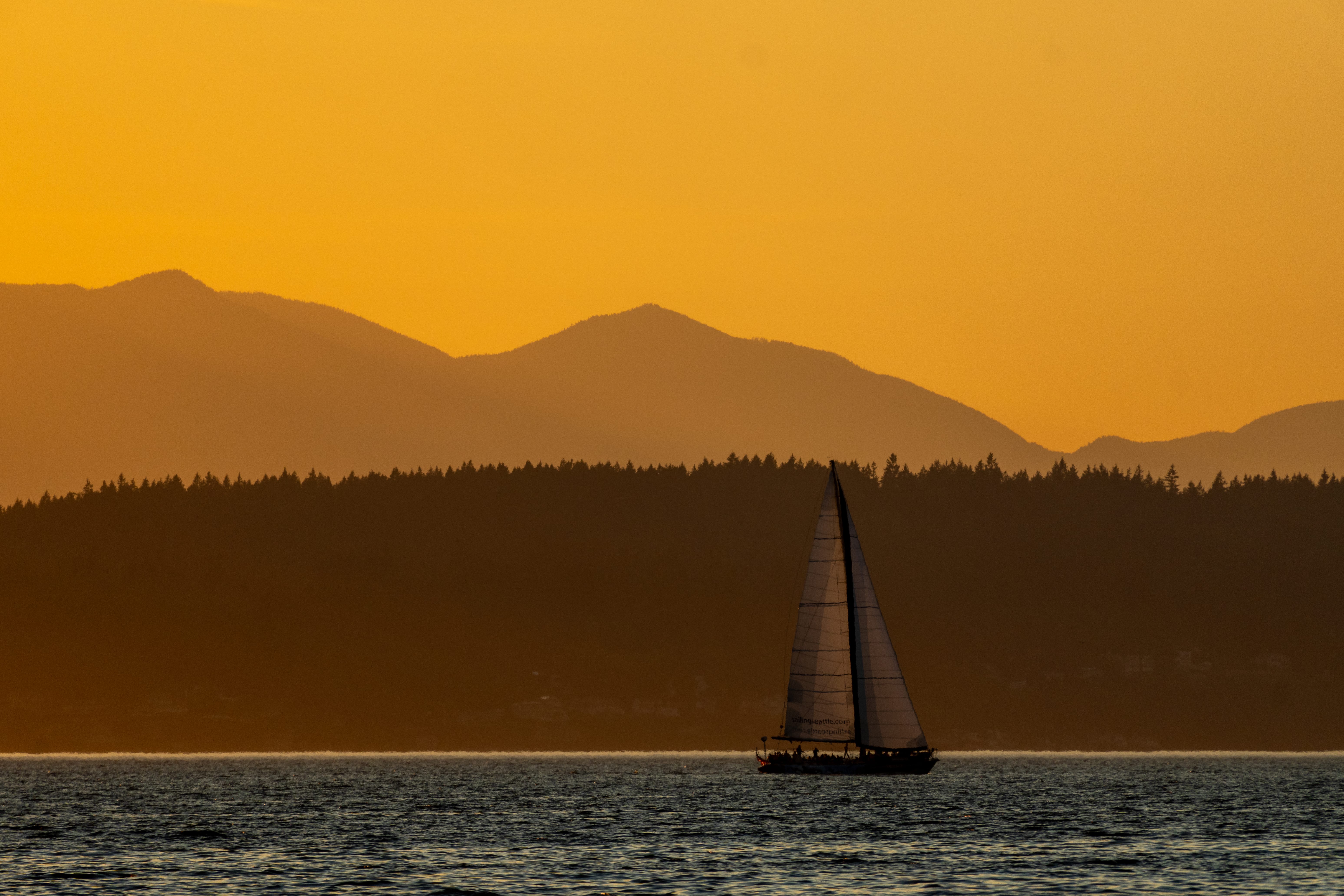 A sailboat crosses dark water before layered mountains and a golden sky.
