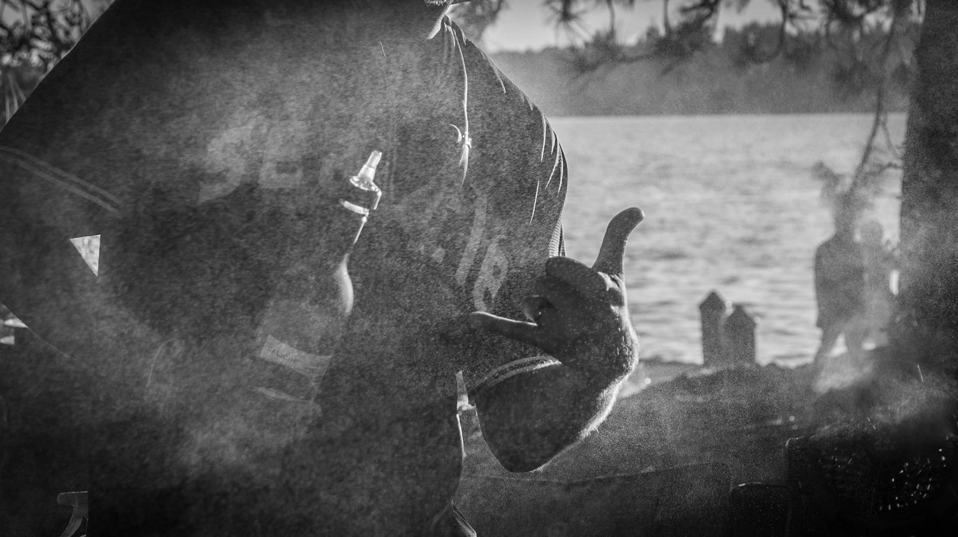 Someone bent over a smoking grill at the water's edge in black and white, figures standing out in the shallows behind.