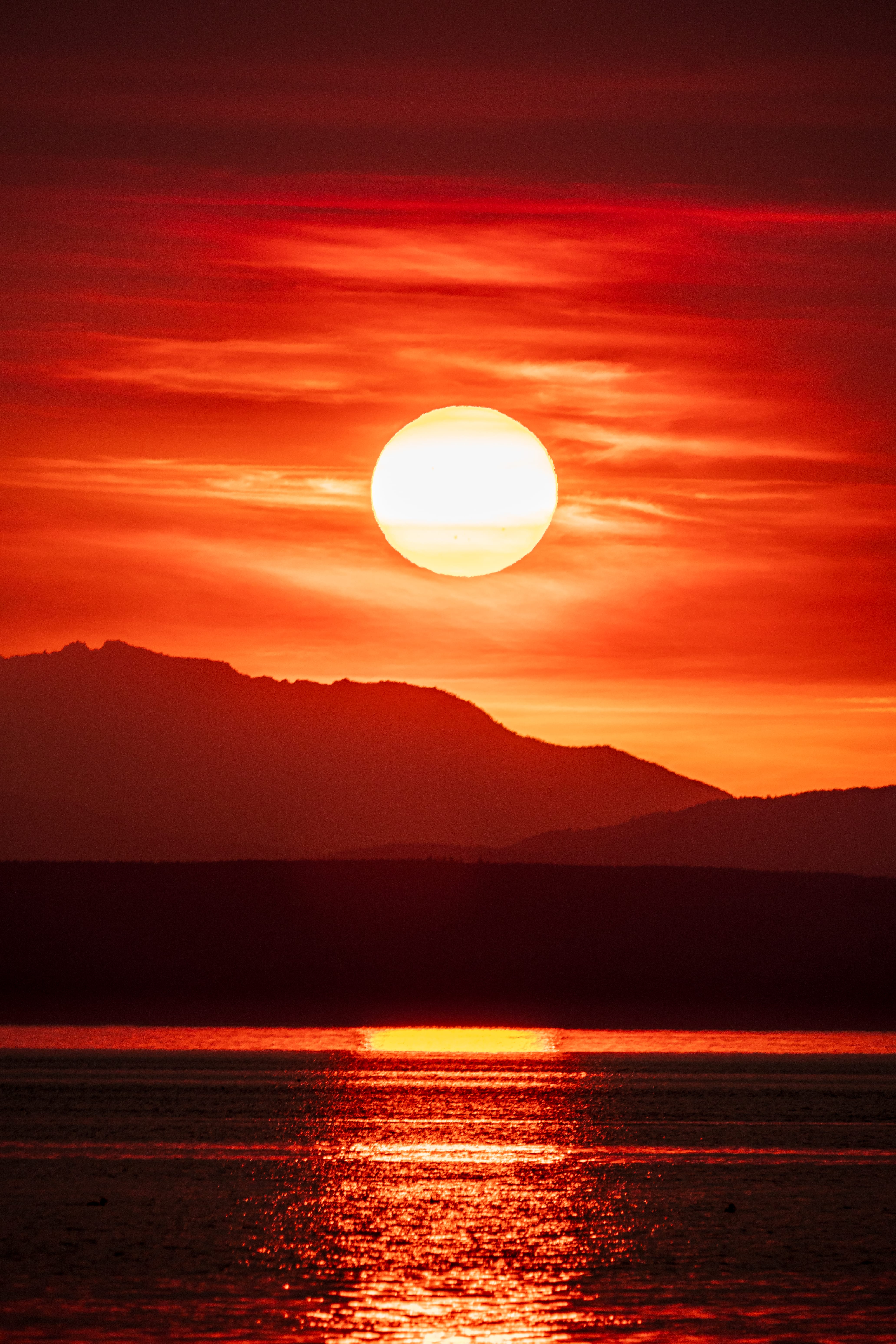 A red sun sets over a dark mountain ridge, reflected in the water below.