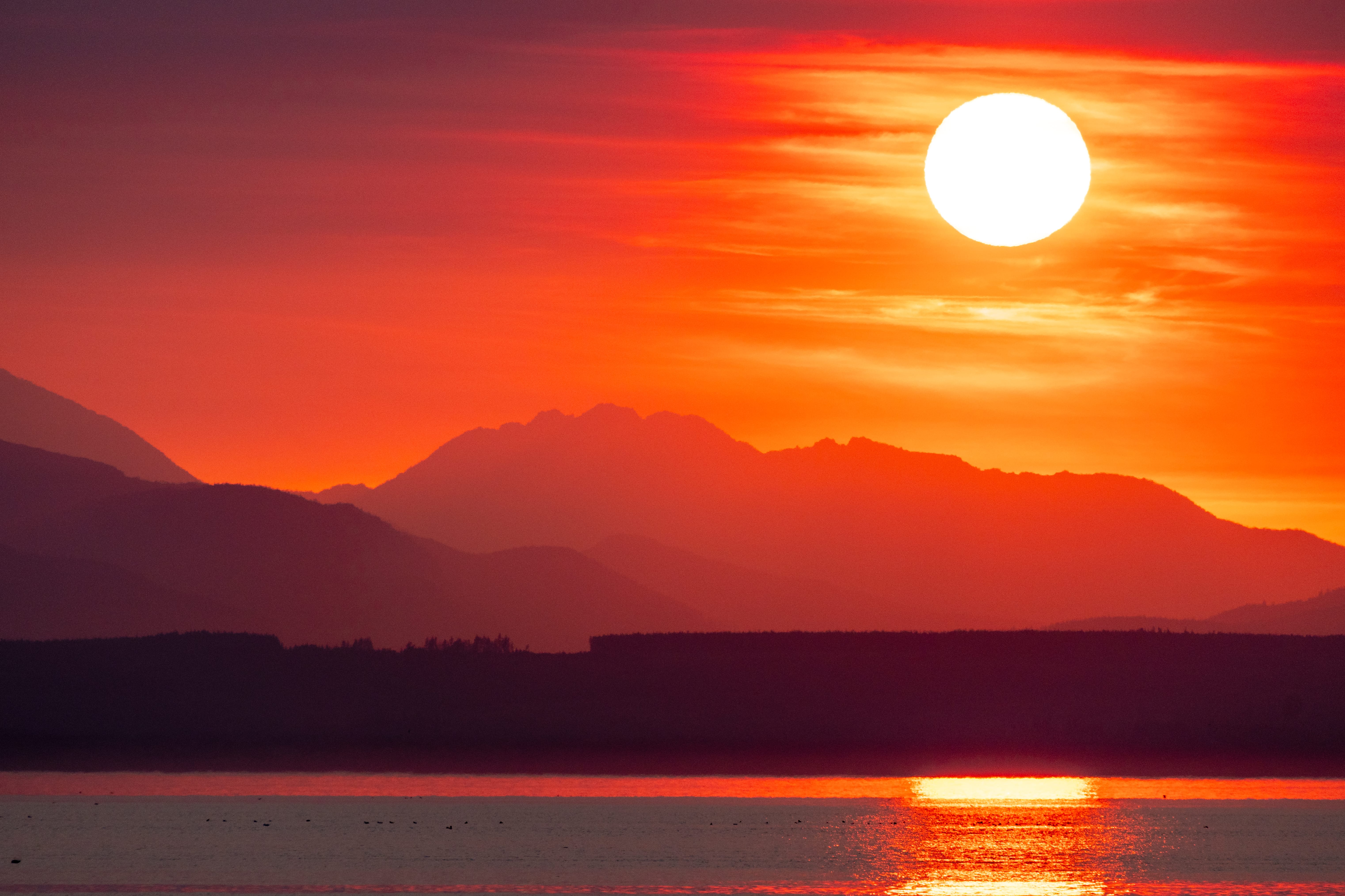 The sun hangs above dark mountain peaks, casting a bright red reflection across the water.