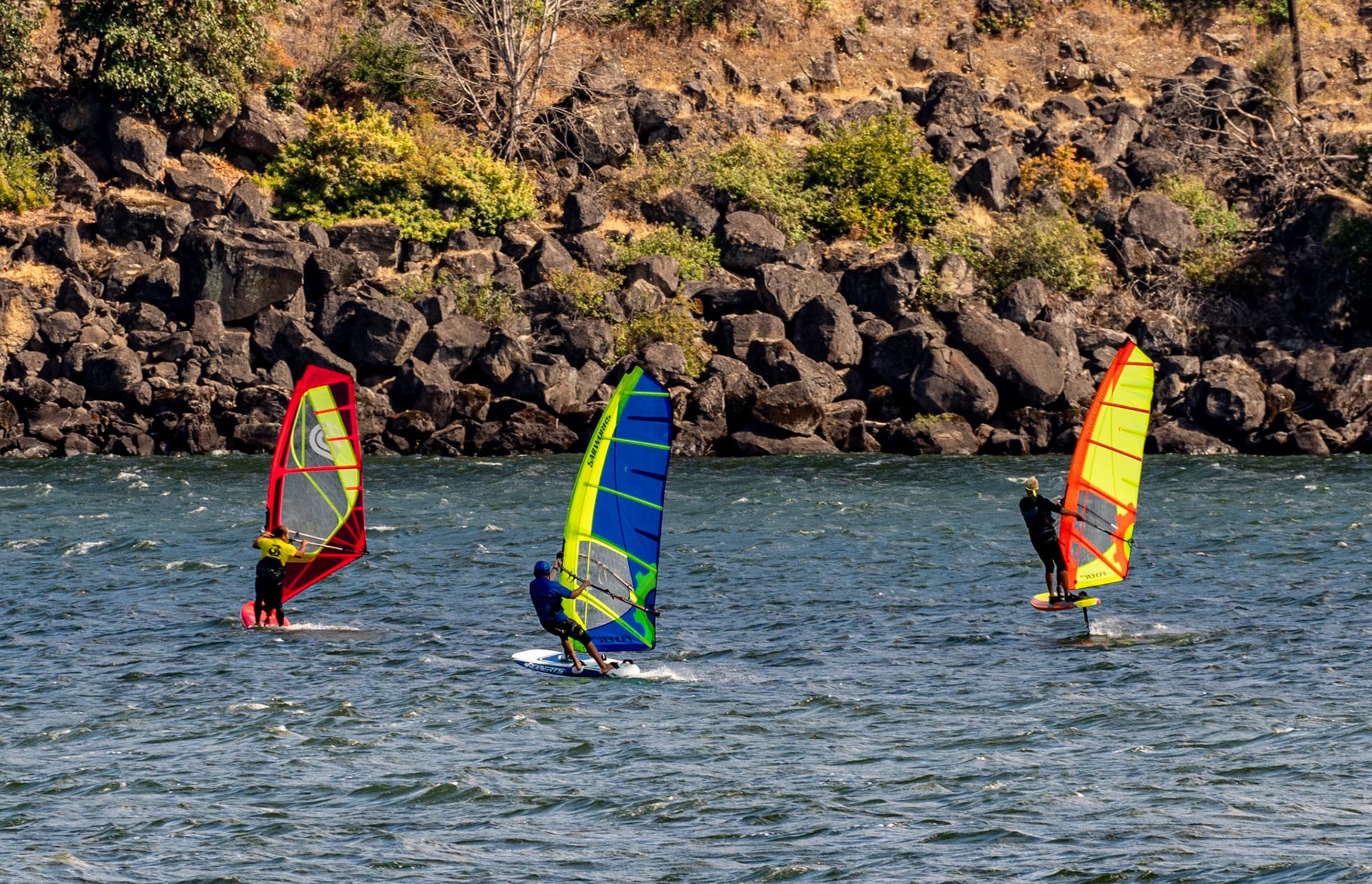 Three windsurfers with colored sails on the river below basalt cliffs.