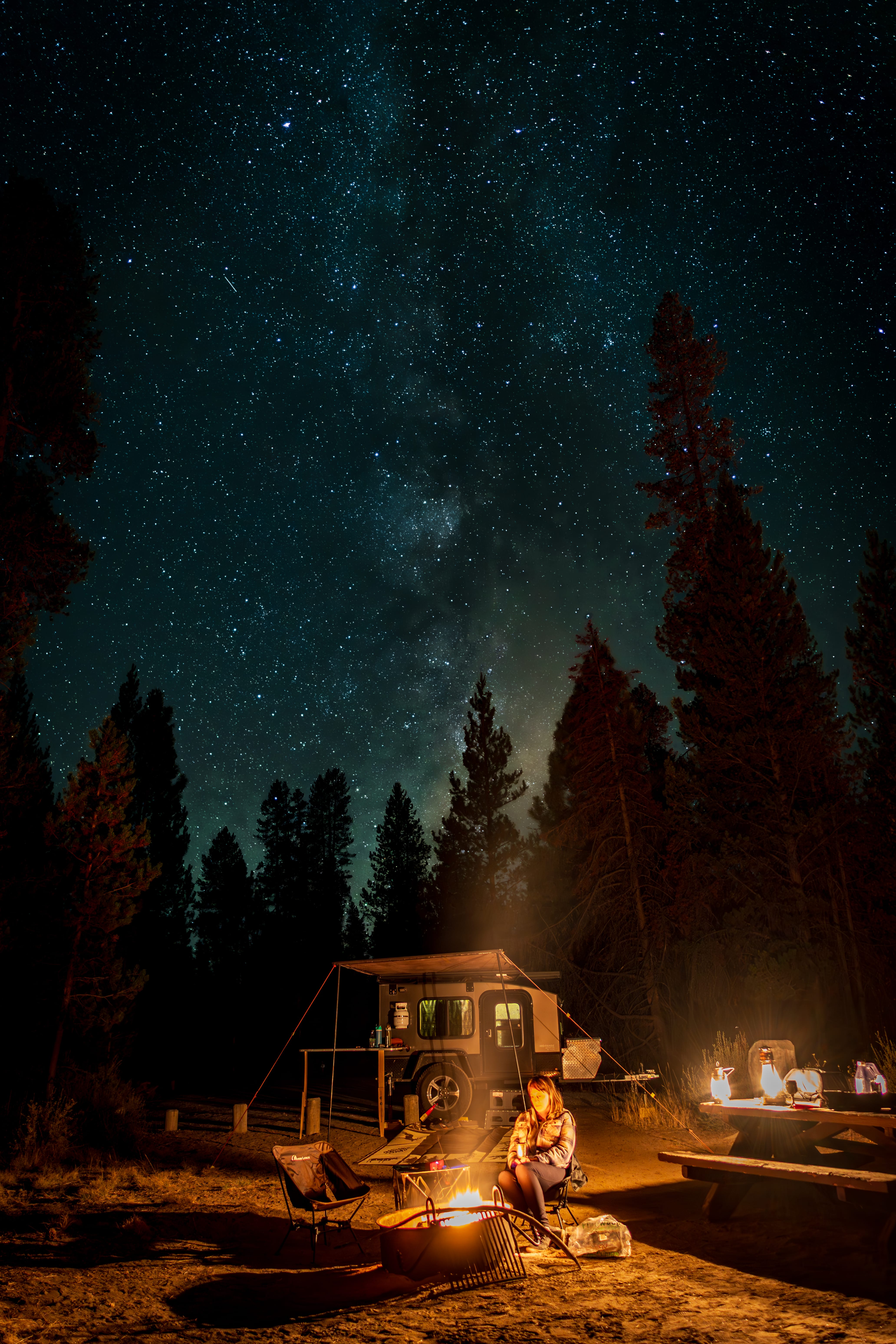 A campsite glows beneath the Milky Way, with tall trees silhouetted against the stars.