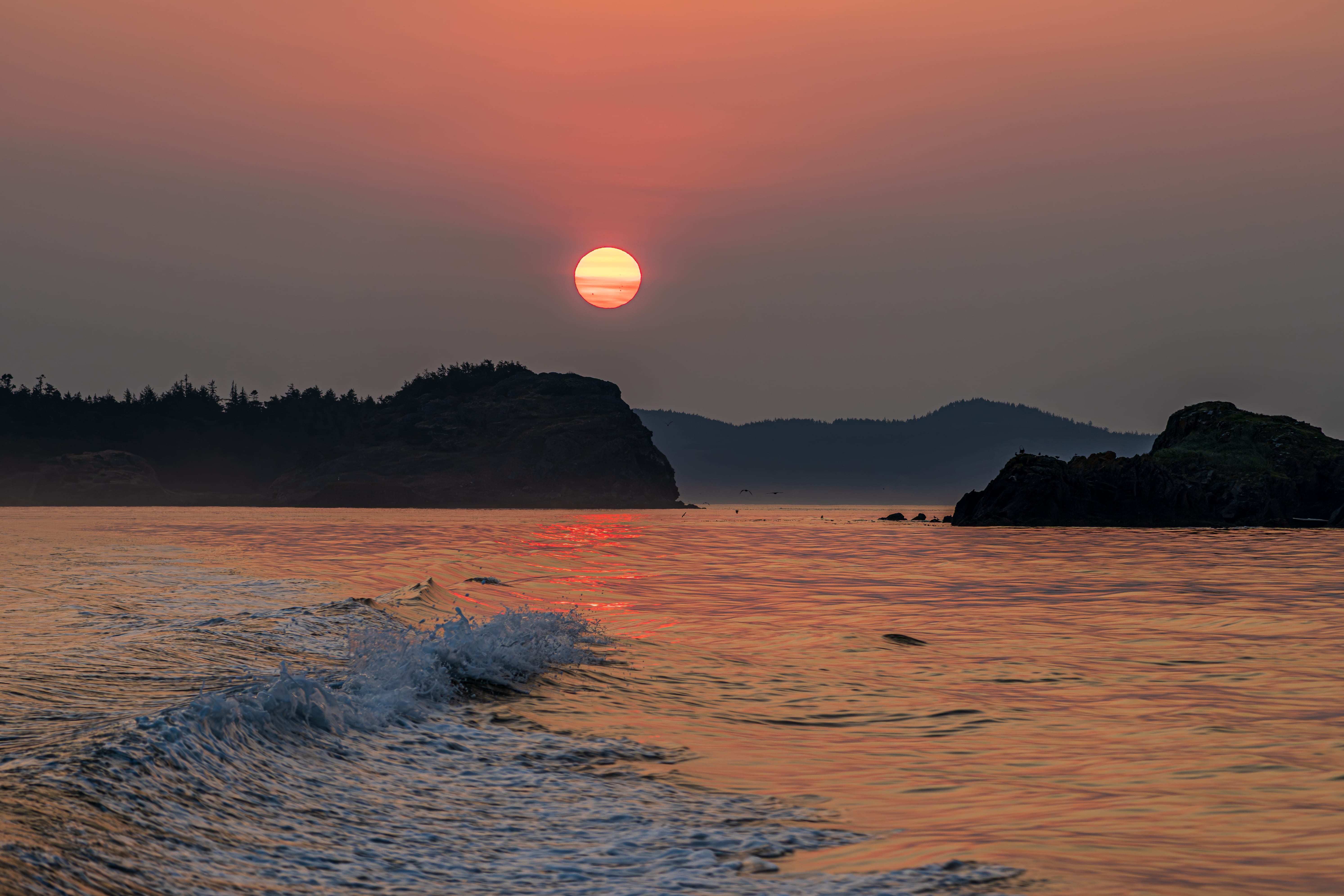 The setting sun glows over low coastal islands as a wave rolls ashore.