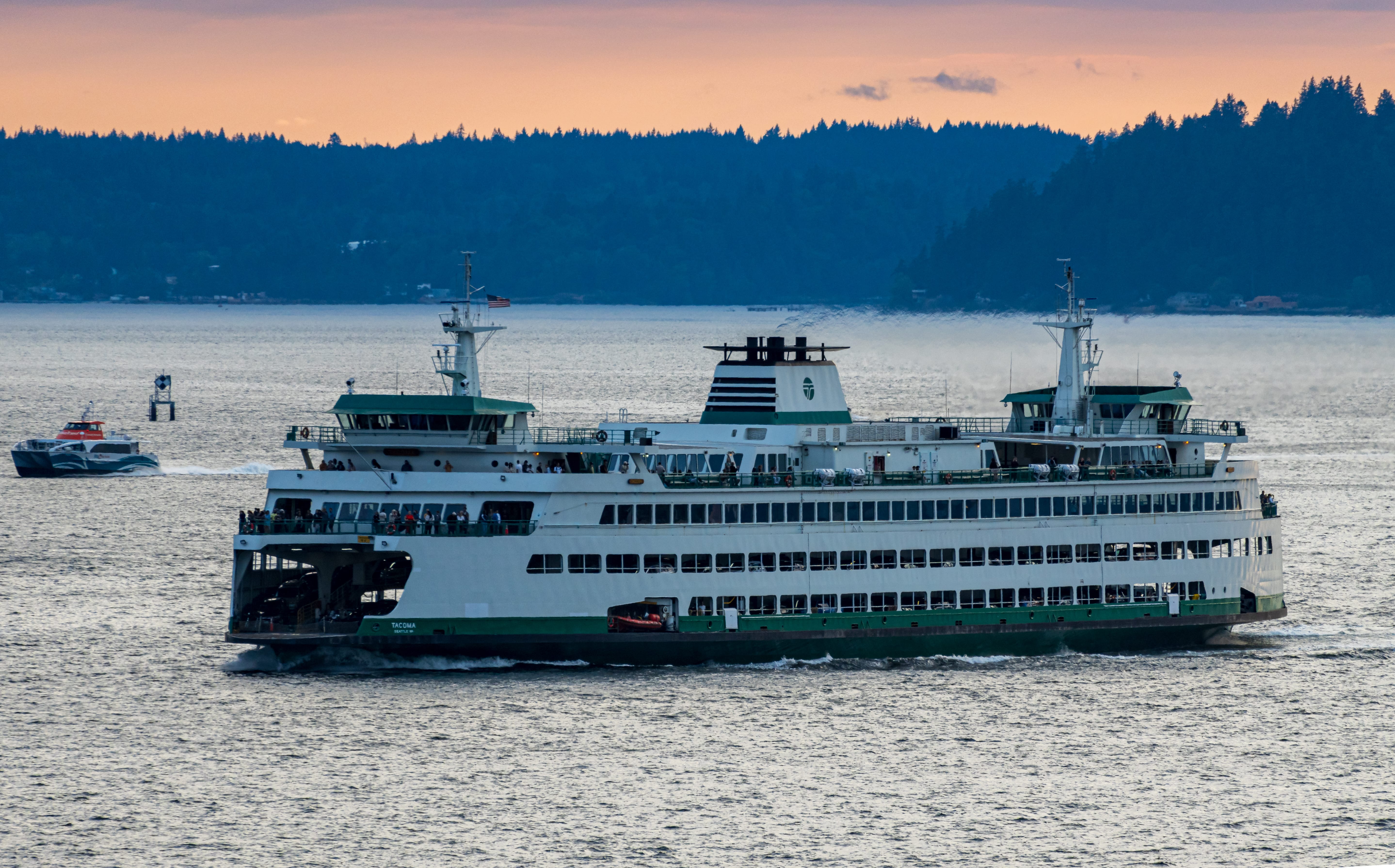 A ferry crosses calm water beneath a pink sunset and dark tree-covered shoreline.