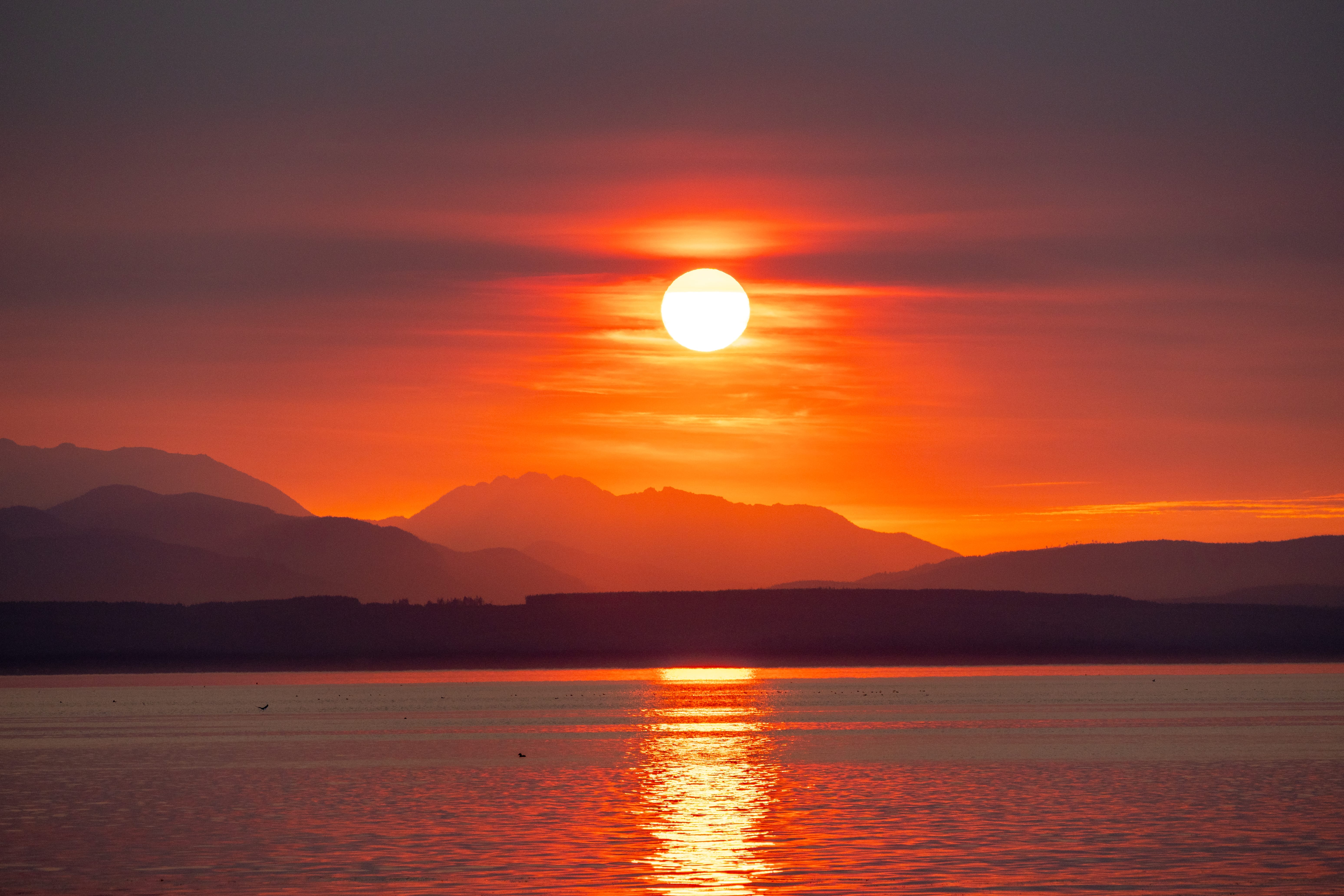 A red-orange sun sets over layered mountain silhouettes and calm water.